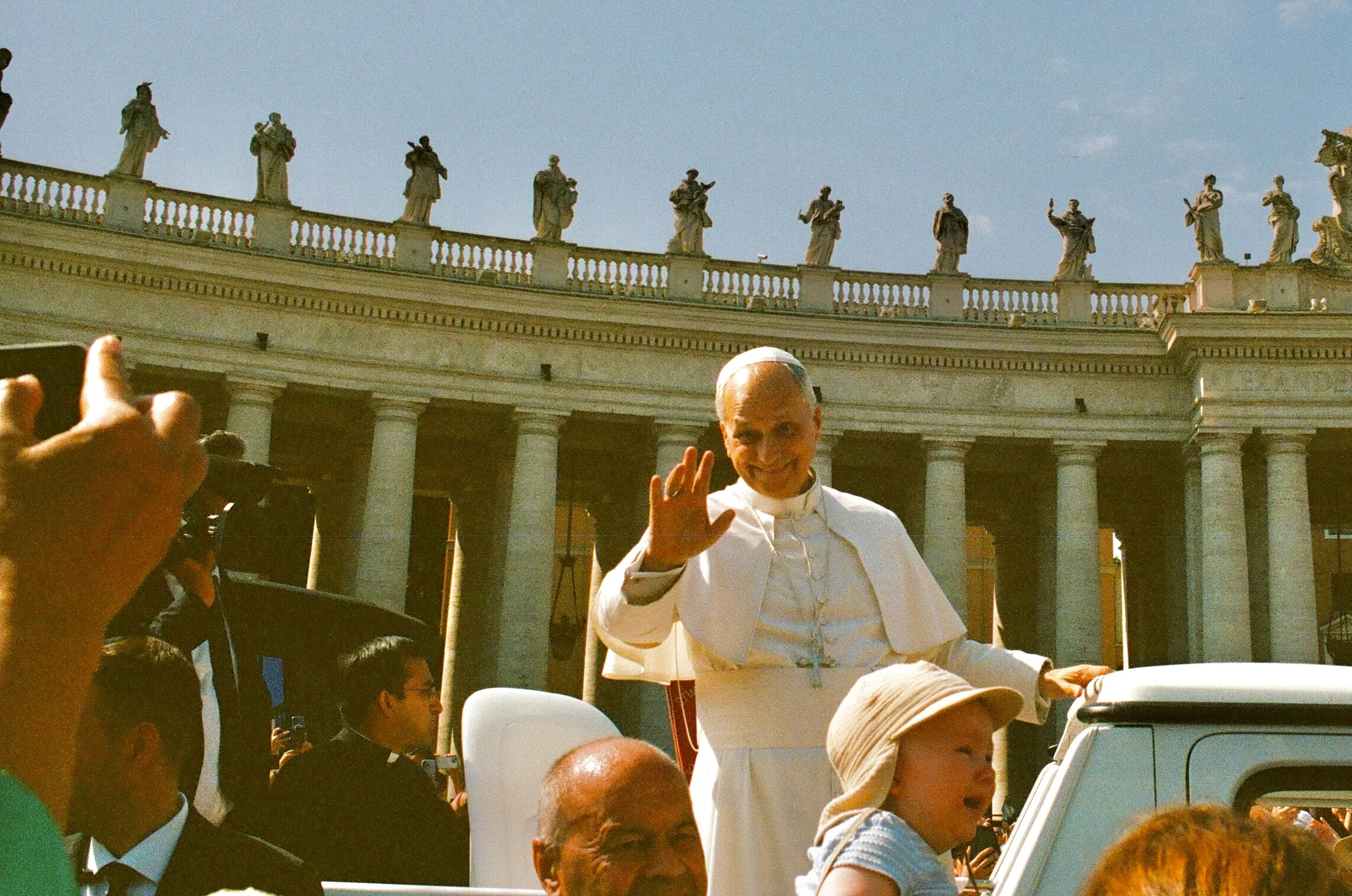 The pope waving from an open vehicle before a crowd and curved colonnade in Rome