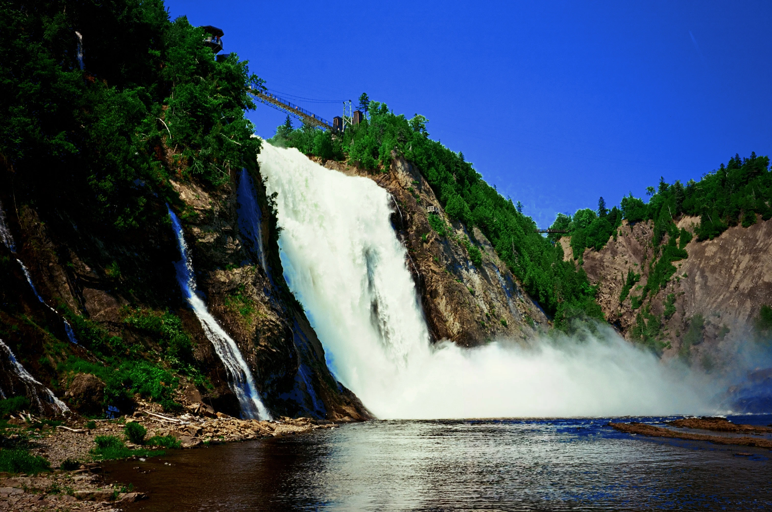 Elliot’s film photograph of a waterfall and suspension bridge, with a cobalt blue sky and deep green trees.
